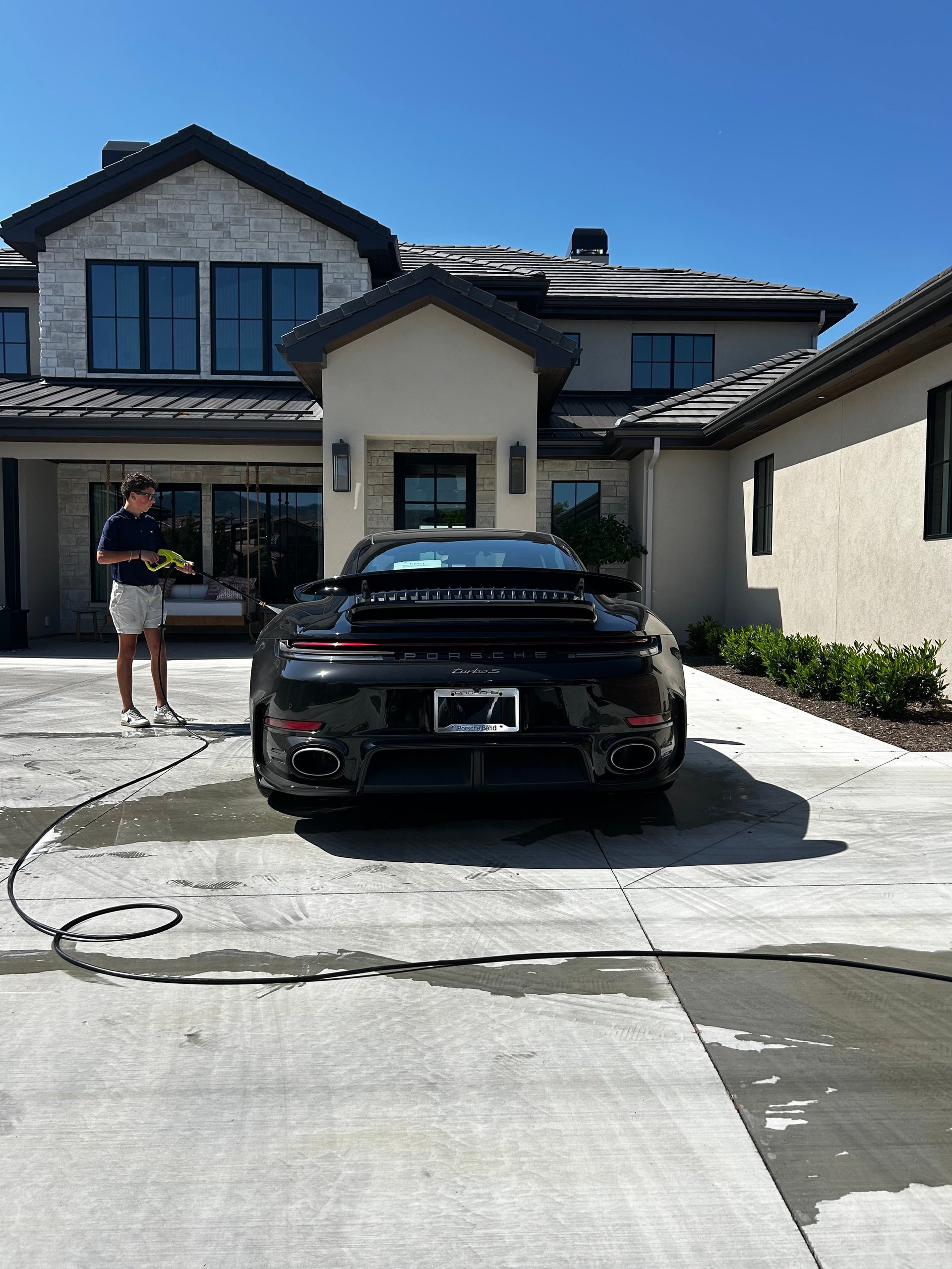 Man washing black sports car in driveway of modern two-story house on sunny day