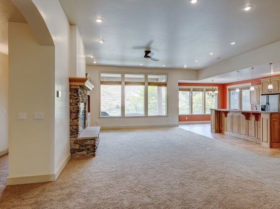 Spacious, modern open-plan living room with carpeted flooring, recessed lighting, fireplace, large windows, and orange cabinetry visible in the kitchen area