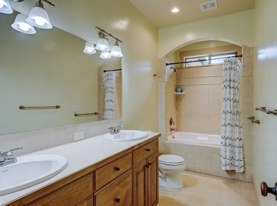 Bright bathroom with pale yellow walls, wooden vanity with double sinks, and bathtub with shower enclosure