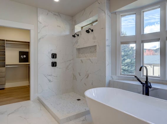 Modern bathroom with marble shower enclosure, freestanding white tub, black faucet, and large windows overlooking landscape