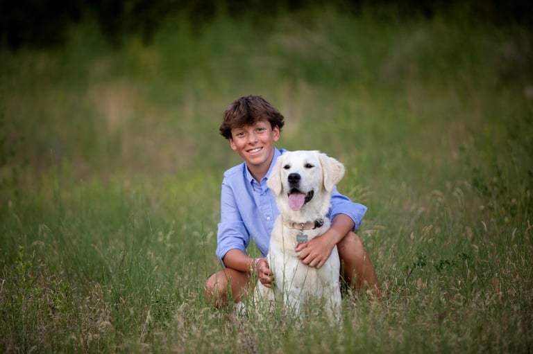 Young boy in blue shirt crouching in grass with white Labrador dog, both smiling at camera
