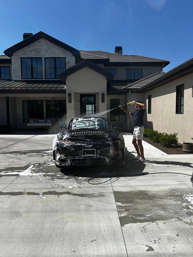 Person washing a black car on a driveway in front of a modern two-story house with white and gray exterior under clear blue sky