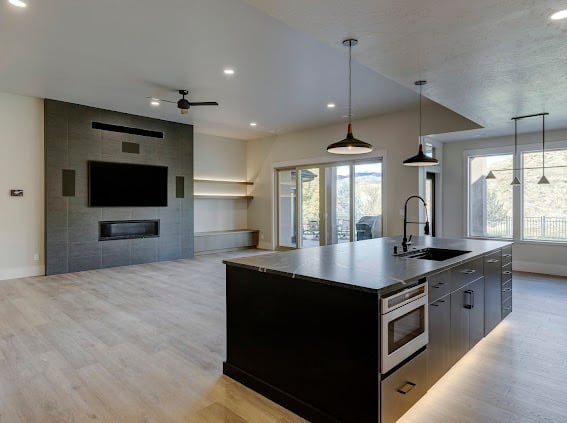 Modern open-concept kitchen and living room with black island, stainless steel appliances, tile fireplace, and natural light from large windows