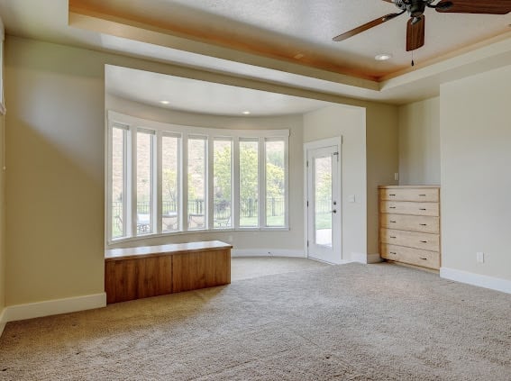 Empty bedroom with large multi-pane windows, ceiling fan, wood trim, and light carpet flooring