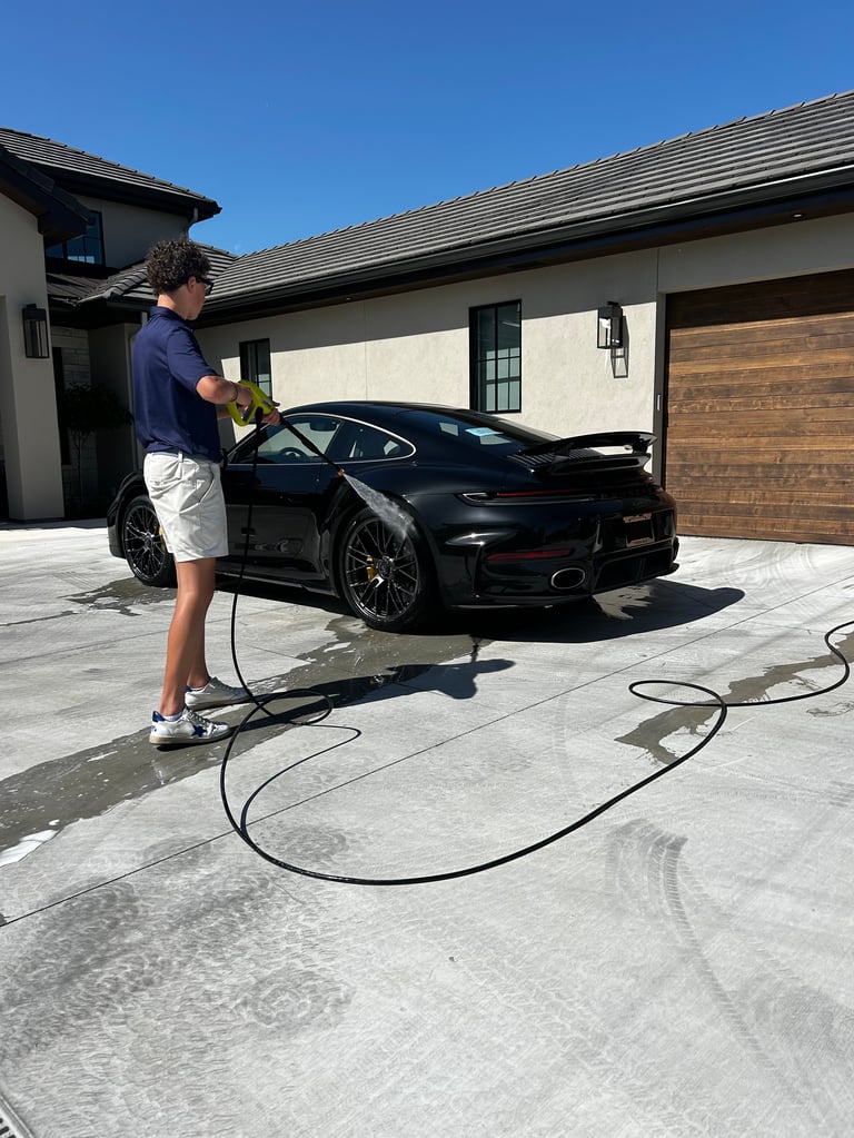 Man washing black sports car with hose on modern garage driveway under clear blue sky
