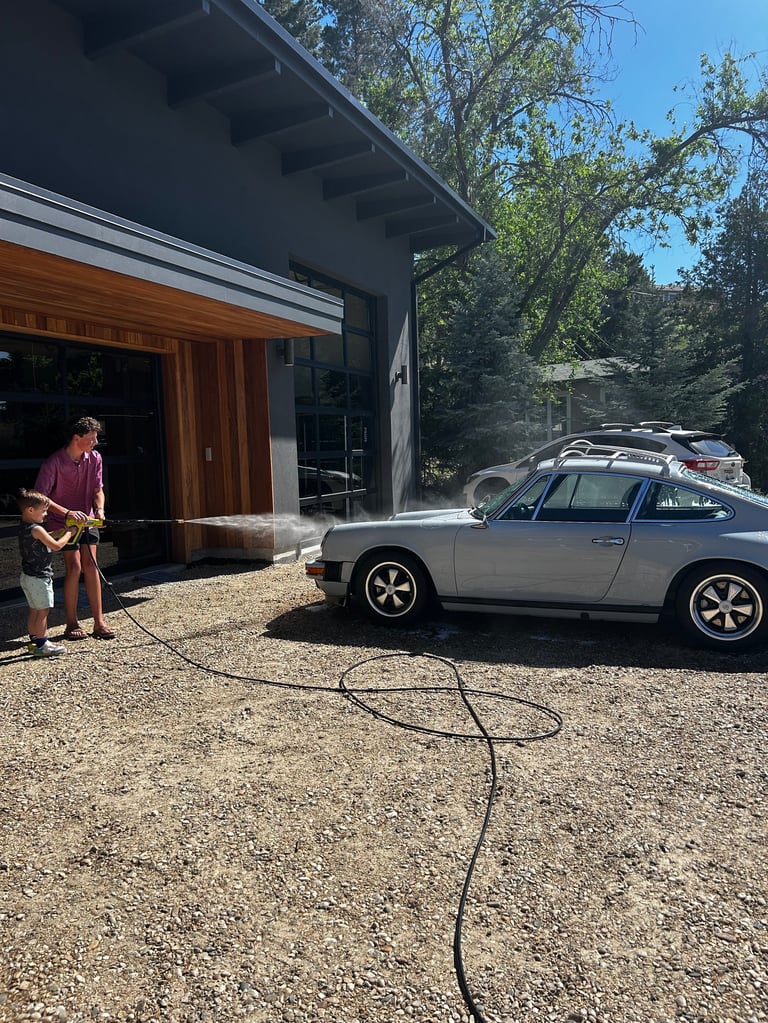 Man with guitar standing beside silver car being washed with hose in front of modern house