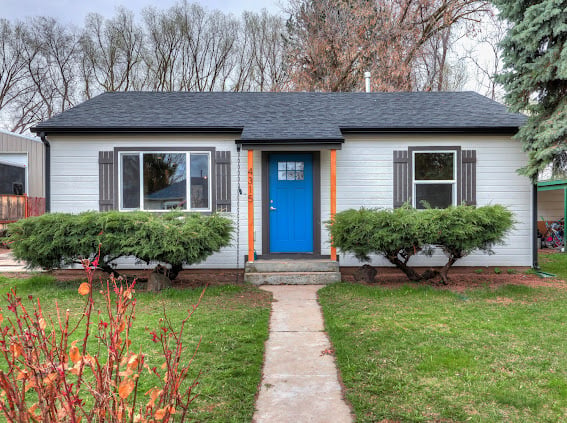 Front view of a single-story white house with blue door, dark roof, landscaped front yard with shrubs and concrete walkway