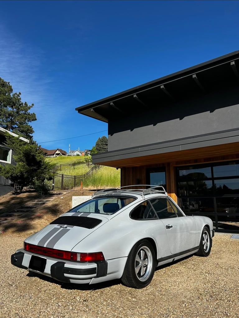 White Porsche 911 with racing stripes parked in front of modern house with black roof and large windows