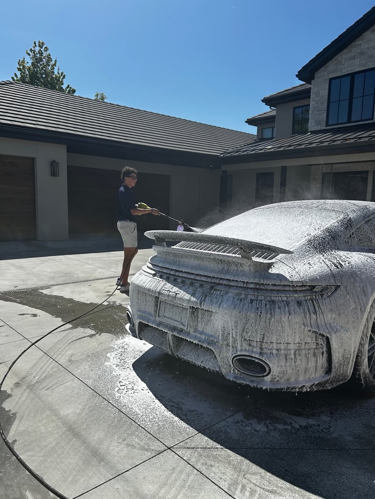 Person washing a soapy luxury car in a modern home driveway on a sunny day