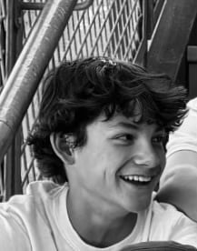 Black and white portrait of a smiling young man with dark wavy hair wearing a light-colored shirt, photographed outdoors
