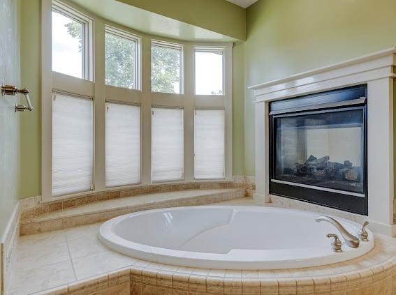 Luxurious spa bathroom with large circular sunken bathtub, wall-mounted flat-screen TV, and bay windows with natural lighting