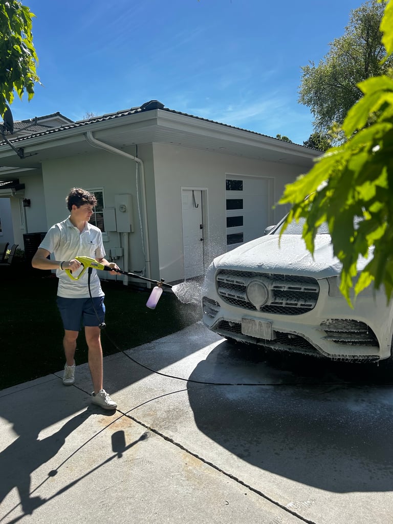 Person washing a white SUV with a hose in a driveway in front of a modern house on a sunny day
